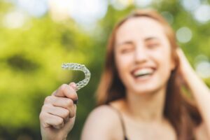 Young woman outside blurry in the background smiling holding Invisalign to foreground